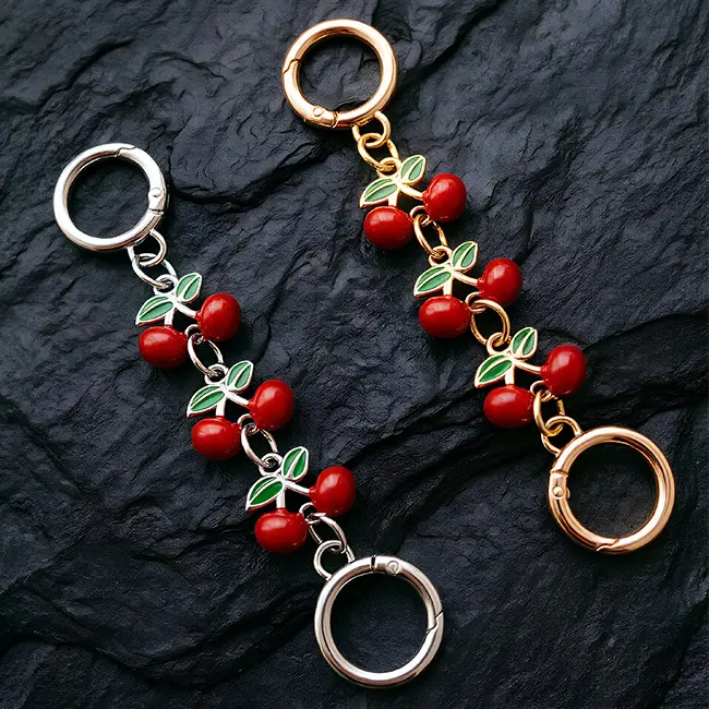 Vista principal de charms de cereza esmaltada mostrando detalles en esmalte rojo y verde sobre cadenas decorativas en aleacion de zinc plata y oro claro