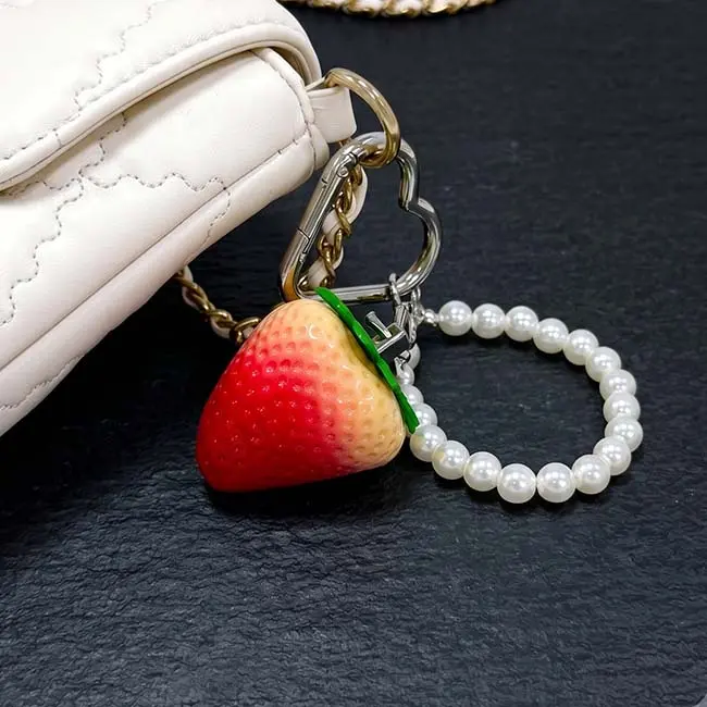 An application showcase of the semi-ripe eco-resin strawberry component utilised as a luxury bag charm secured to the polished metal chain hardware of a pristine white quilted designer handbag accessory.