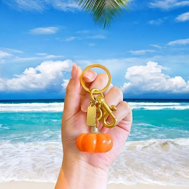 A handheld scale reference view of the glossy orange eco-resin pumpkin keyring held in a hand against a scenic tropical ocean beach backdrop demonstrating real-world accessory sizing and weight.