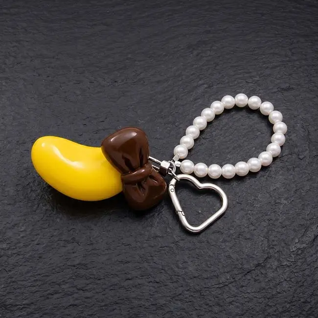 A detailed macro view of the yellow eco resin banana charm with a rich chocolate brown bowknot showing its glossy reflective surface durable silver toned metal connections and a loop of lustrous faux pearls on slate.