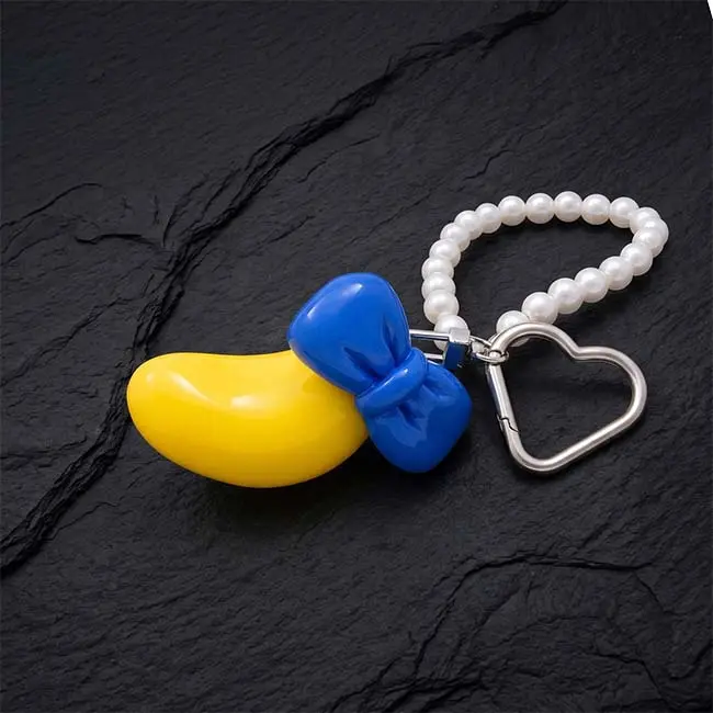 A premium high gloss yellow eco resin banana keyring featuring a vibrant blue bowknot accent paired with a polished silver zinc alloy heart shaped keyring clasp and a decorative white faux pearl loop strand on slate.