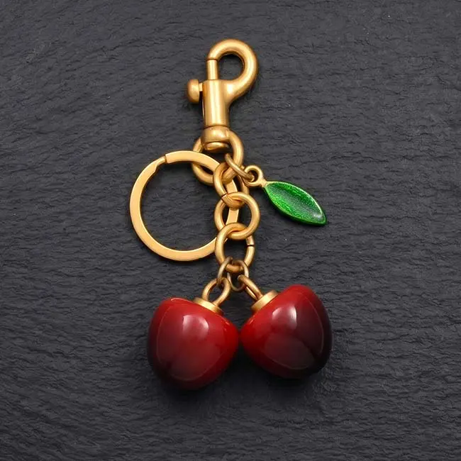 A detailed close up view of the dual red eco resin cherry charm showing the polished reflective finish sturdy golden zinc alloy metal loop connections and a vibrant green leaf element on slate.