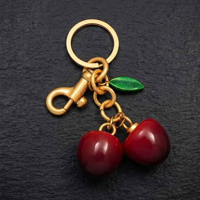 A premium high gloss red eco resin twin cherry keyring component featuring a vibrant green leaf accent paired with a durable gold toned zinc alloy spring snap hook and split keyring on slate.