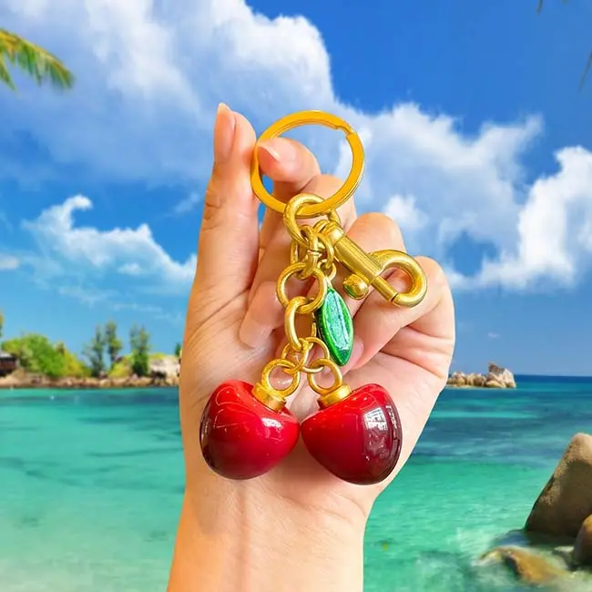 A handheld scale reference view of the glossy red eco resin cherry keyring component held in a hand against a scenic tropical ocean beach backdrop demonstrating real world item size and weight.