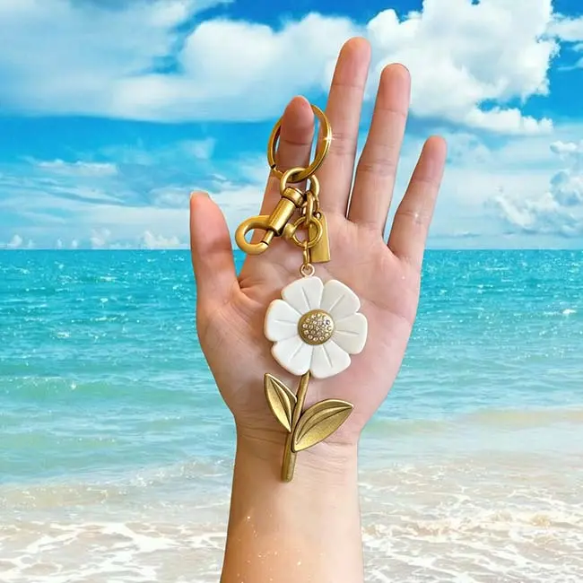 مقارنة الحجم في اليد لميدالية مفاتيح الزهرة البيضاء من الراتنج الصلب غير الشفاف باللون الذهبي العتيق أمام خلفية شاطئ استوائي