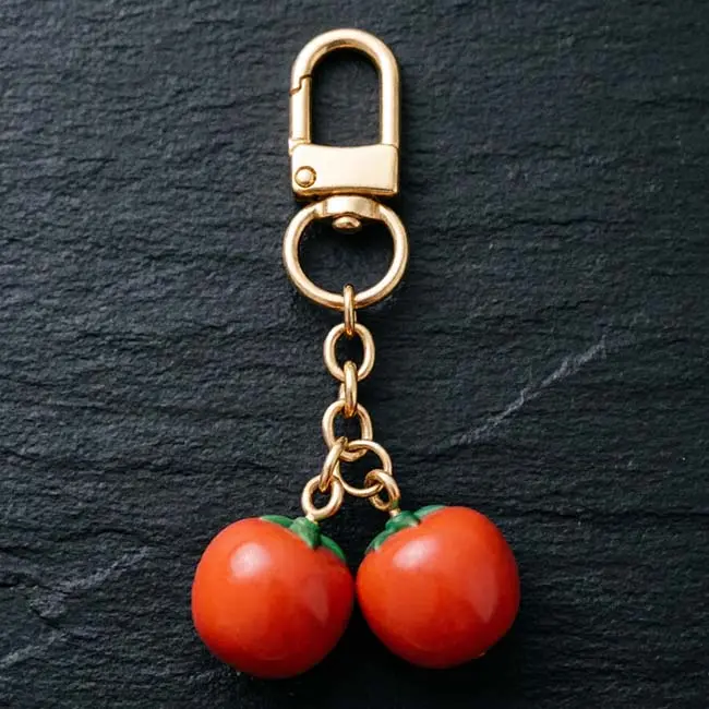 A premium solid opaque resin hardware twin persimmon keychain component featuring glossy vibrant orange fruit pendants with green leafy tops, attached to a durable gold toned zinc alloy spring snap hook.