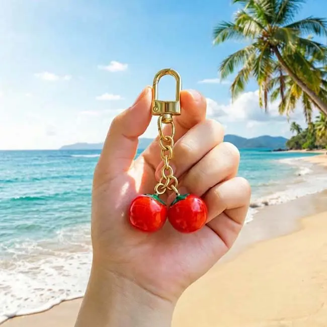 A handheld scale reference view of the solid opaque resin hardware persimmons keychain held in a hand against a beautiful tropical ocean beach backdrop, displaying real world luxury accessory proportions.