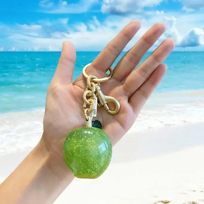 مقارنة الحجم في اليد لميدالية مفاتيح التفاح الأخضر الكبير من الراتنج البيئي أمام خلفية شاطئ استوائي لتوضيح الأبعاد الحقيقية