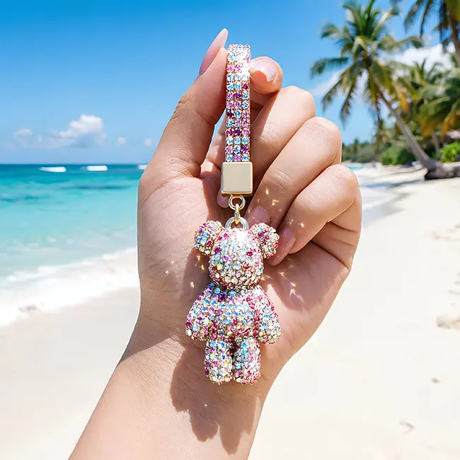 Handheld scale reference view of the pink with iridescent clear factory direct pavé charms featuring a full body optical rhinestone inlaid bear held in a hand against a tropical ocean beach backdrop.