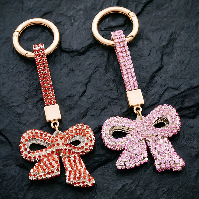 A pair of ruby red and soft pink factory direct pavé charms featuring full body optical rhinestone inlaid bow motifs with matching crystal studded straps and gold hardware on a slate background.