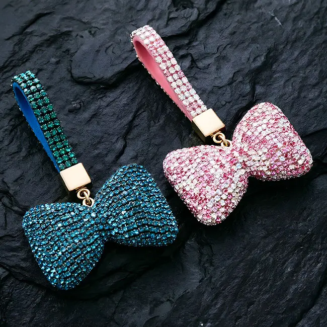Two vibrant factory direct pavé charms featuring full body optical rhinestone inlaid puffy bow motifs in ocean blue and pink with iridescent clear with matching crystal studded straps on a textured slate background.