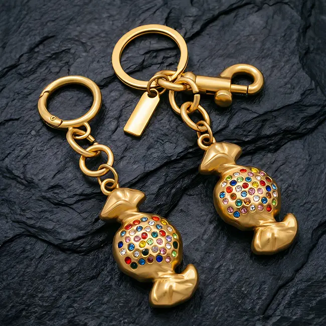 A pair of matte deep gold wholesale rhinestone keychains featuring a boutique candy motif inlaid with random multi-color crystals, two distinct spring hook round spring ring & swivel snap hook