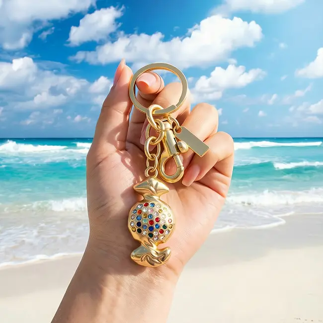 Handheld scale reference view of light champagne gold boutique candy wholesale rhinestone keychains held in a hand against a tropical ocean beach backdrop displaying real world luxury bag charm dimensions.
