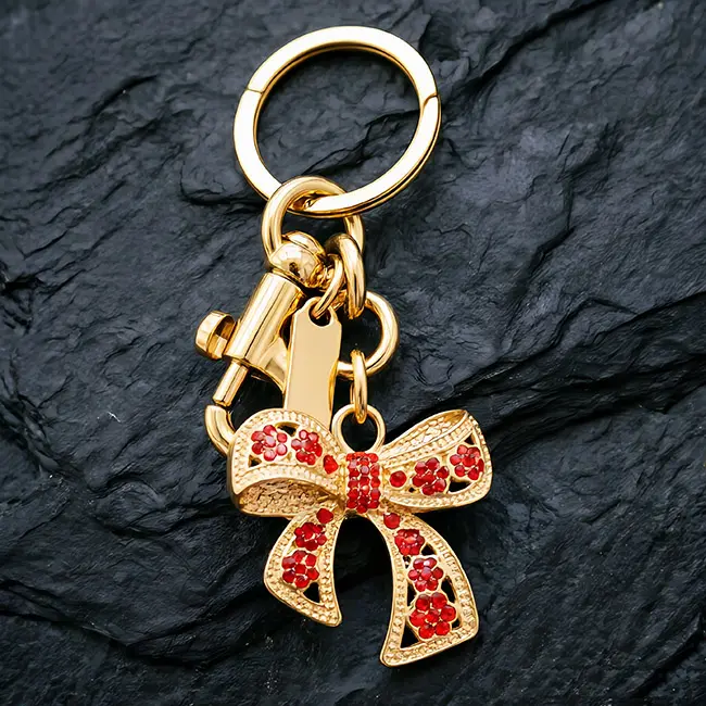 A premium light metallic gold hollow bow custom crystal inlaid components accessory featuring vibrant ruby red optical rhinestones, a heavy-duty swivel snap hook, and a blank metal tag on a dark slate.