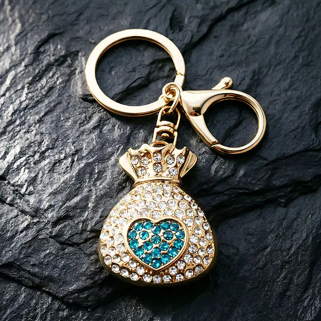 A macro material detail view of the B2B optical crystal molding accessory highlighting a teal blue rhinestone heart on the fortune bag charm with a robust zinc alloy clasp on a dark slate mat.