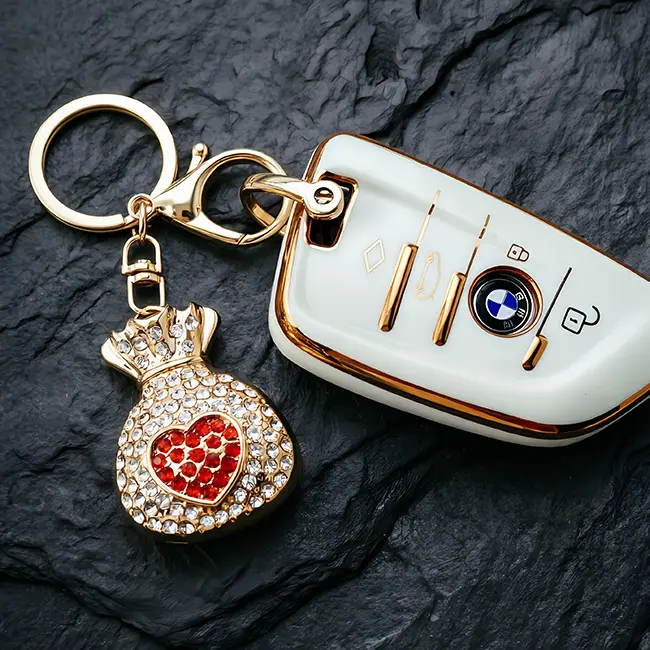 A scale reference view of the B2B optical crystal molding accessory featuring a vibrant ruby red rhinestone fortune bag charm placed beside a modern white luxury car key to display real world size.