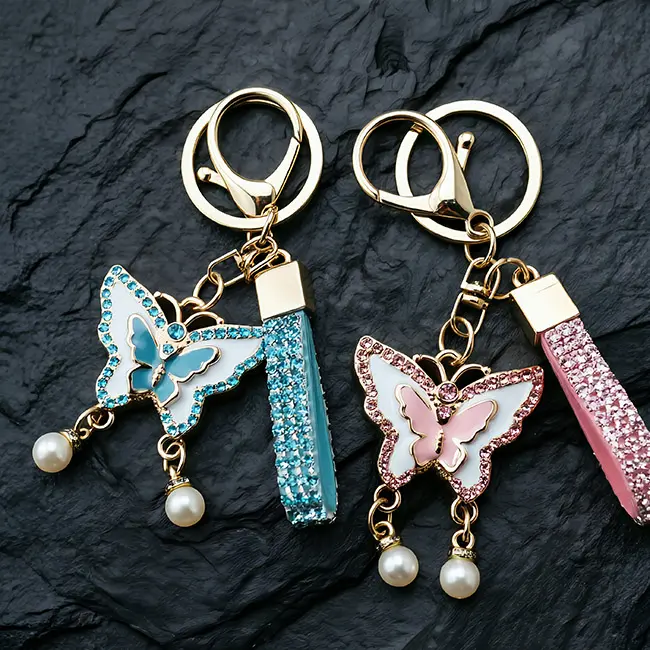 A pair of premium light gold wholesale rhinestone keychains featuring elegant butterfly charms with teal blue and soft pink enamel infill, matching crystal straps, and delicate pearl dangles.