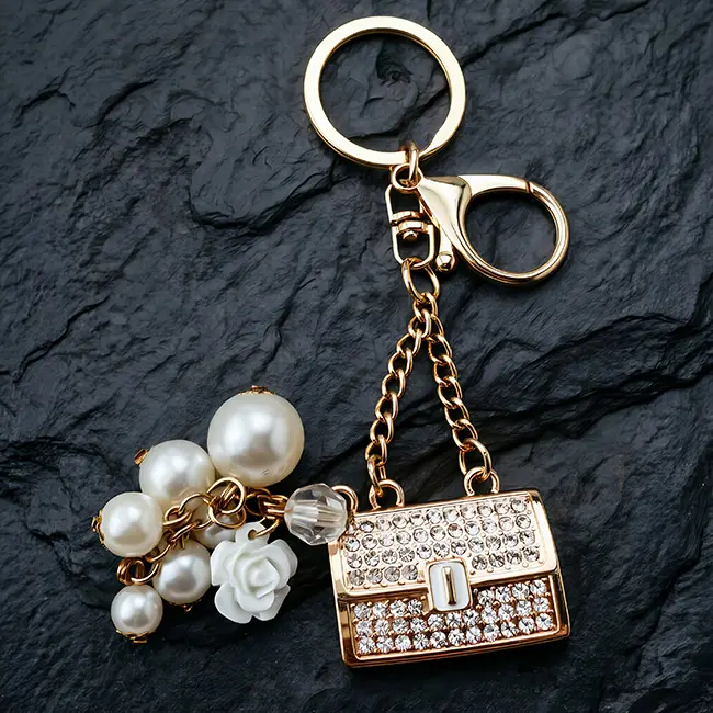 B2B optical crystal molding keychain showcasing a crystal clear rhinestone enamel fortune purse with lustrous pearl dangles and a robust gold clasp on a dark mat.