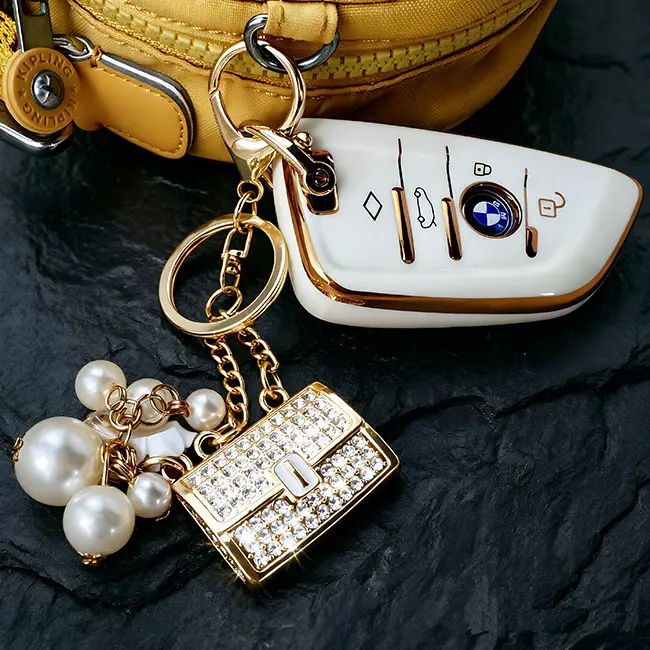 A scale reference view of the B2B optical crystal molding accessory featuring a crystal clear rhinestone enamel fortune purse charm and pearl dangles placed next to a modern white luxury vehicle key.