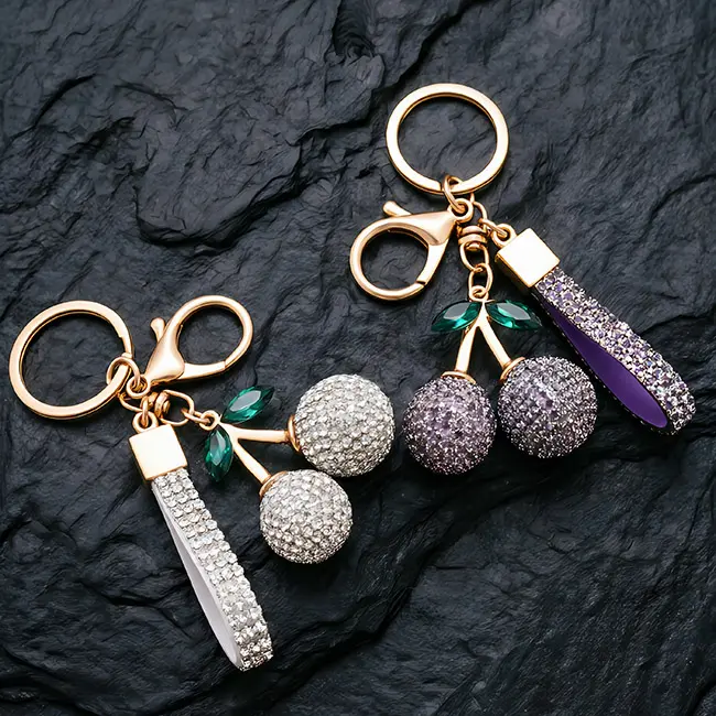 A macro detail view of the light gold factory direct pavé charms showcasing crystal clear and dusty mauve (purple-pink) optical rhinestone cherries with robust zinc alloy hardware and green leaf accents.