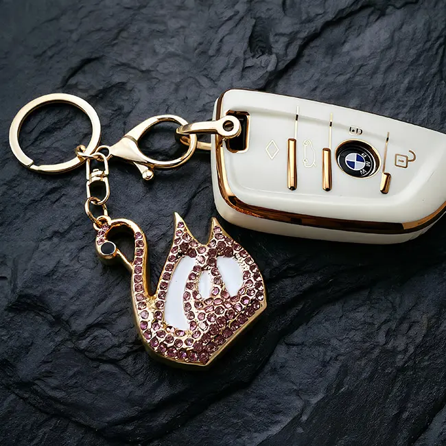 A scale reference view of the B2B optical crystal molding accessory featuring a soft pink rhinestone goose charm with crisp white enamel infill placed next to a modern luxury white vehicle key.