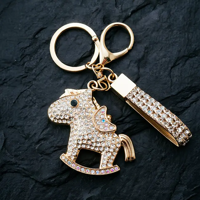 A macro material detail view of the bulk crystal hardware keychain showcasing a crystal clear iridescent clear rhinestone rocker horse carrying a gold ingot with a matching studded strap on a slate background.