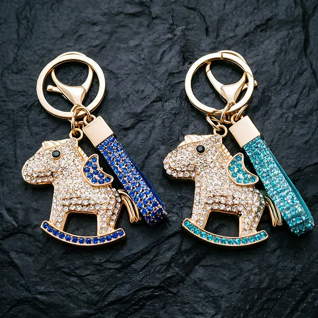 A detailed view of the bulk crystal hardware showcasing two rocker horse charms in cobalt blue and teal blue optical rhinestones, featuring light gold zinc alloy plating on a dark textured mat.