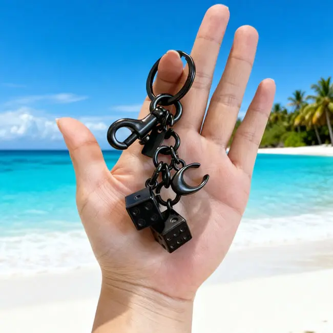 Handheld scale reference view of the matte black precision die casting keyrings dice accessory held in a hand against a tropical ocean beach backdrop displaying real world luxury bag charm dimensions.