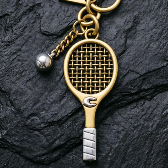 A macro material detail view of the antique finish metal casting keyrings showcasing the precision die-cast tennis racket strings - silver tone tennis ball - uppercase C detail - and vintage brass plating.