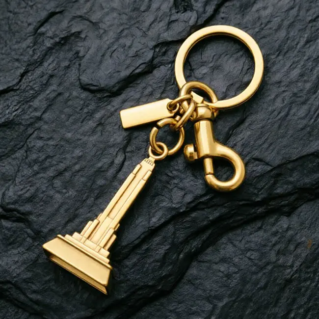 A premium gold bulk 3D moulded charms iron tower component featuring a solid zinc alloy skyscraper - a heavy-duty spring snap hook - and a blank custom logo tag on a textured dark slate background.