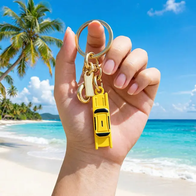 Handheld scale reference view of the yellow bulk 3D moulded enamel infill charms taxi accessory held in a hand against a tropical ocean beach backdrop displaying real world luxury bag charm and keyring sizing.
