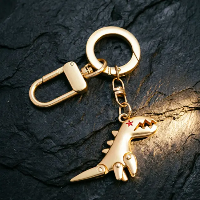 A premium gold dinosaur wholesale Precision Zinc Alloy Castings component featuring a solid metal T-Rex charm with a red star eye - a heavy-duty spring snap hook - and durable metallic hardware links on slate.