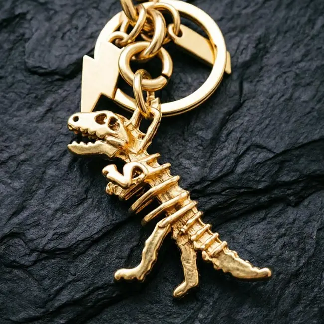 A macro material detail view of the gold Precision Zinc Alloy 3D moulded charms keyring showcasing the precision die-cast dinosaur skeleton ribs - skull structure - and gold electroplated finish.