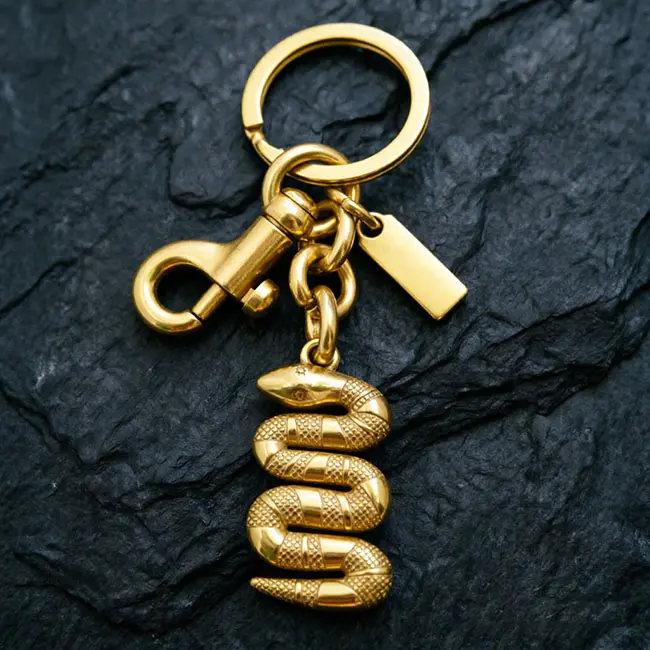A premium electroplated gold snake Precision Zinc Alloy 3D moulded charms component featuring a detailed coiled serpent charm - a heavy-duty spring snap hook - and a blank custom logo tag on slate.